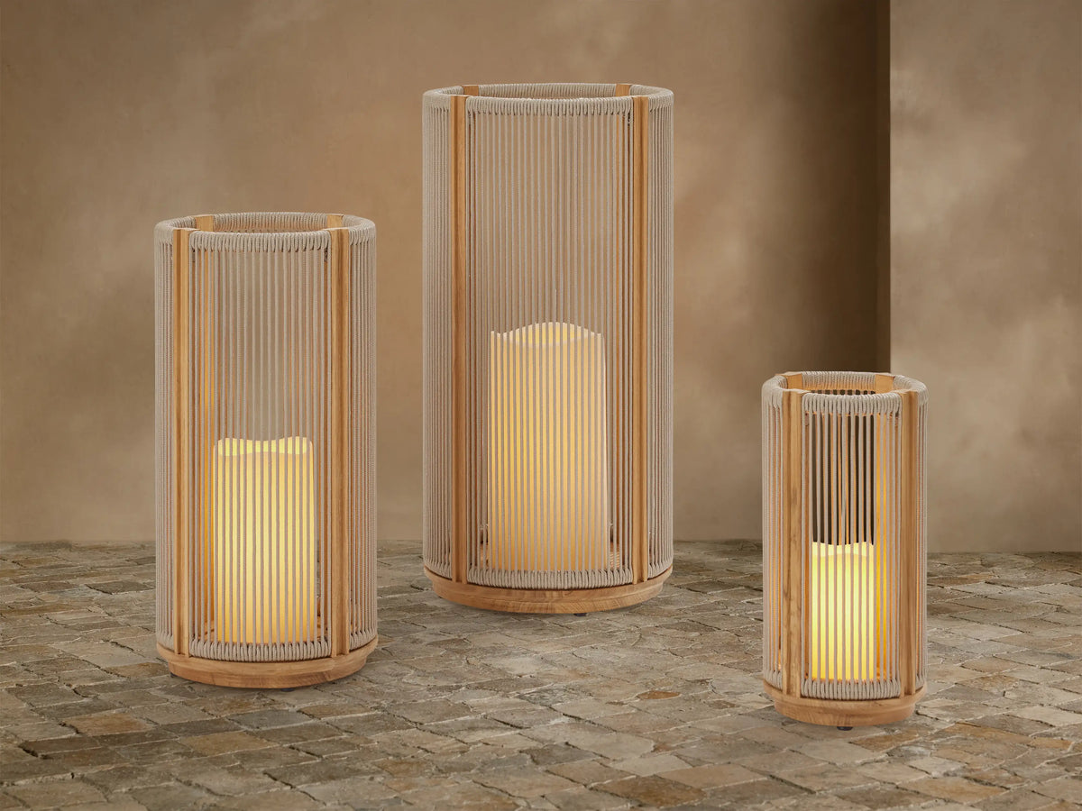 Three Ravello Outdoor Lanterns of varying sizes with natural teak frames and woven beige rope, each glowing with a flameless candle, on a stone patio.