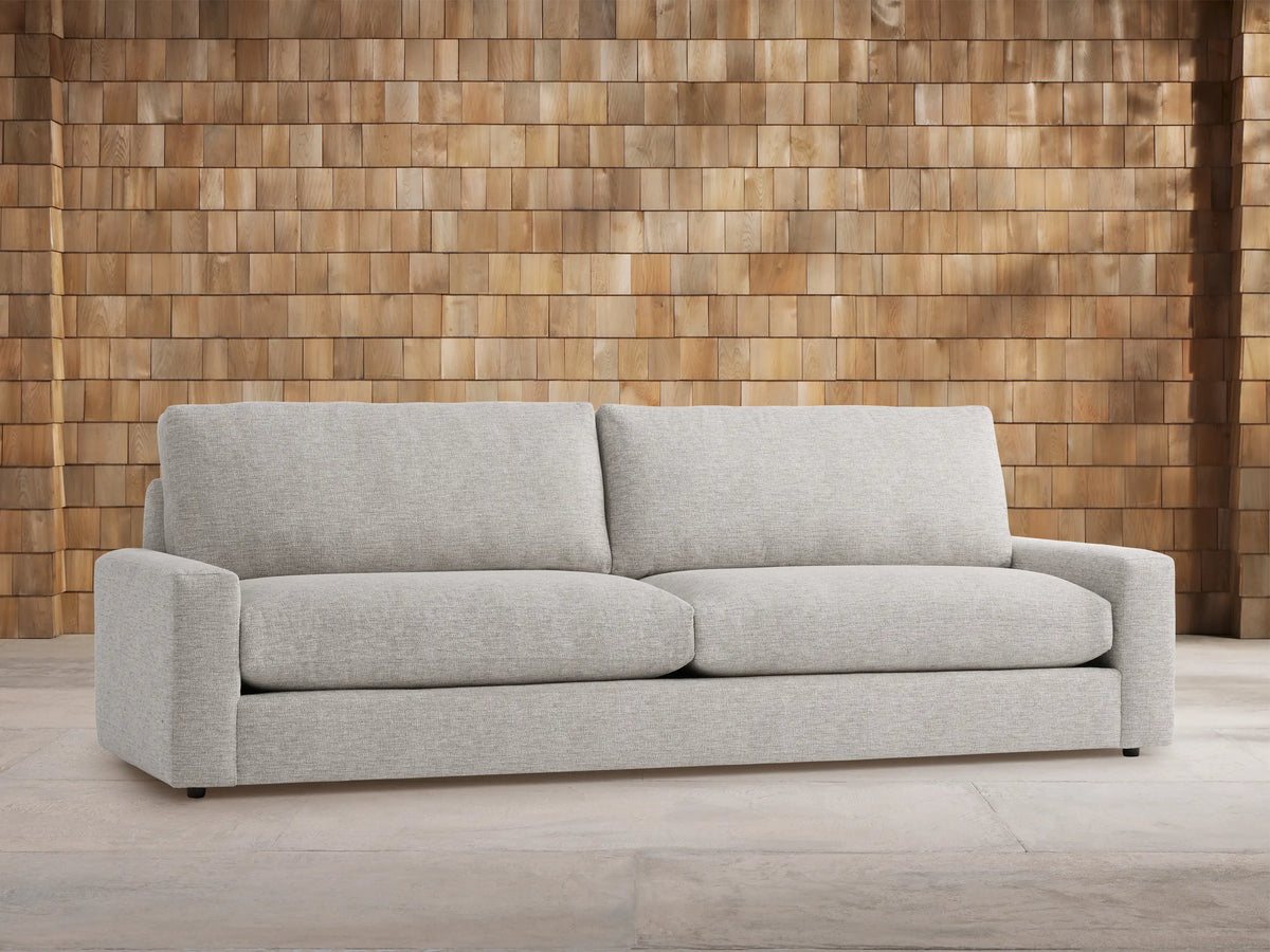 Light gray Remington Outdoor Sofa, front view. Plush two-seater with wide arms and cushions, against a warm wooden shingle wall.