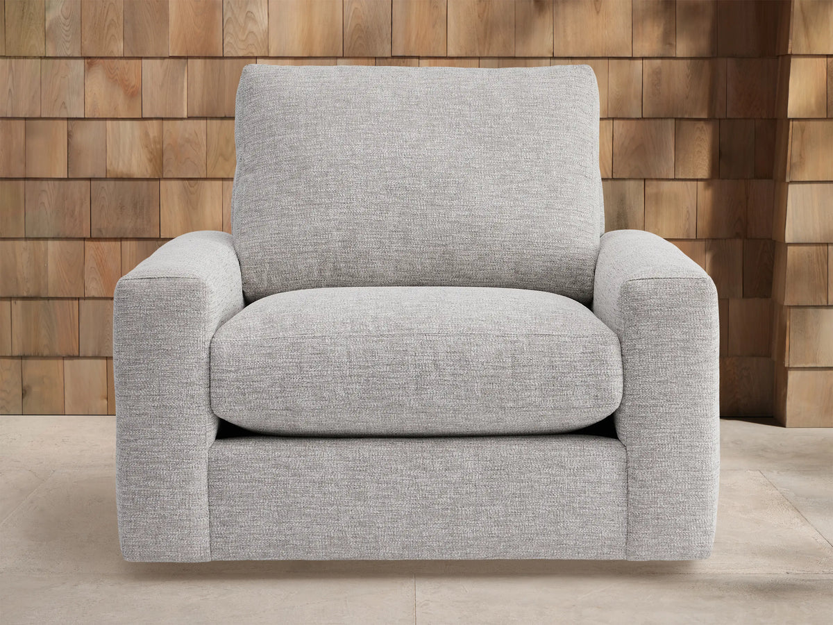 Light grey Remington Outdoor Swivel Chair with plush cushions, featuring square arms, set against a wooden shingle wall background.