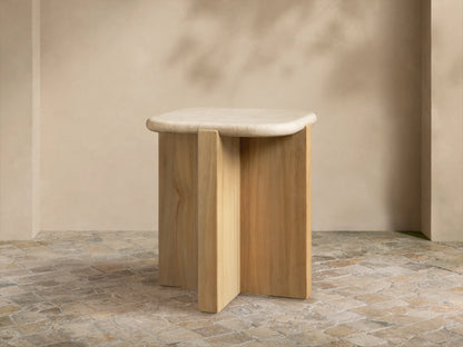 Reily Outdoor End Table with a light marble top & natural teak wood cross-base on stone tiles against a beige wall.
