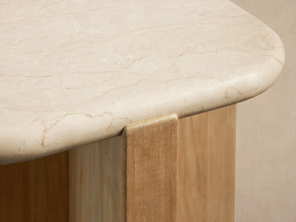 Reily Outdoor End Table with a light marble top & natural teak wood cross-base on stone tiles against a beige wall.