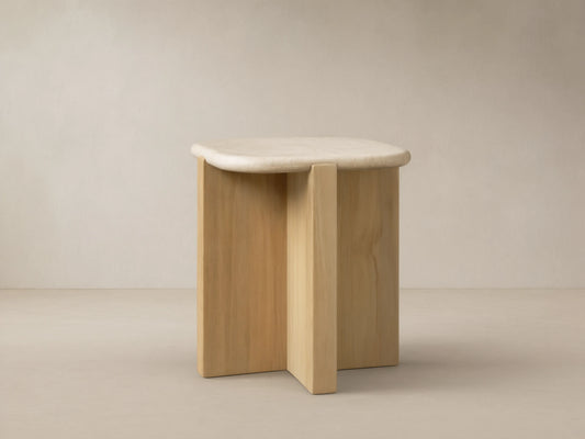 Reily Outdoor End Table with a light marble top & natural teak wood cross-base on stone tiles against a beige wall.