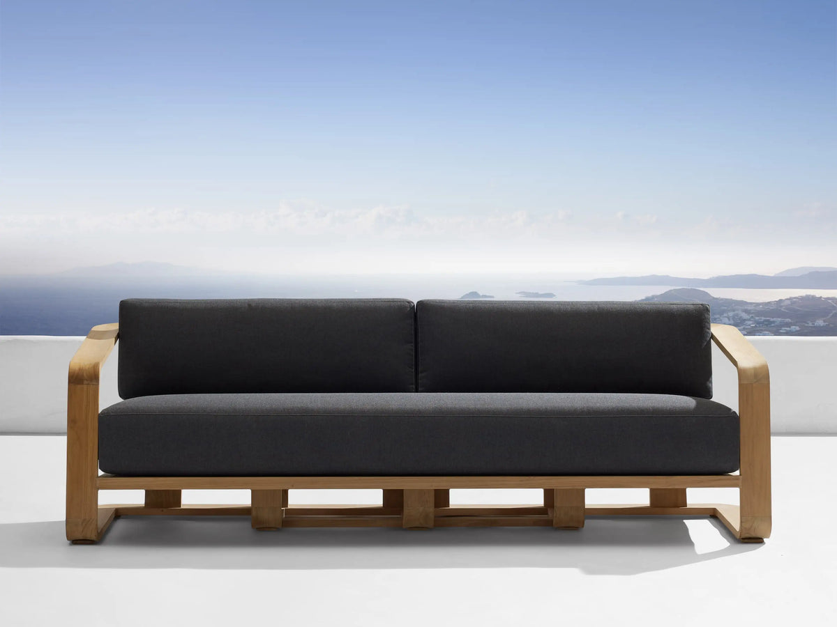 Dark gray Sonoma Outdoor Water-Repellent Replacement Cushions on a wooden sofa, overlooking blue ocean & sky.