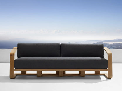 Dark gray Sonoma Outdoor Water-Repellent Replacement Cushions on a wooden sofa, overlooking blue ocean & sky.