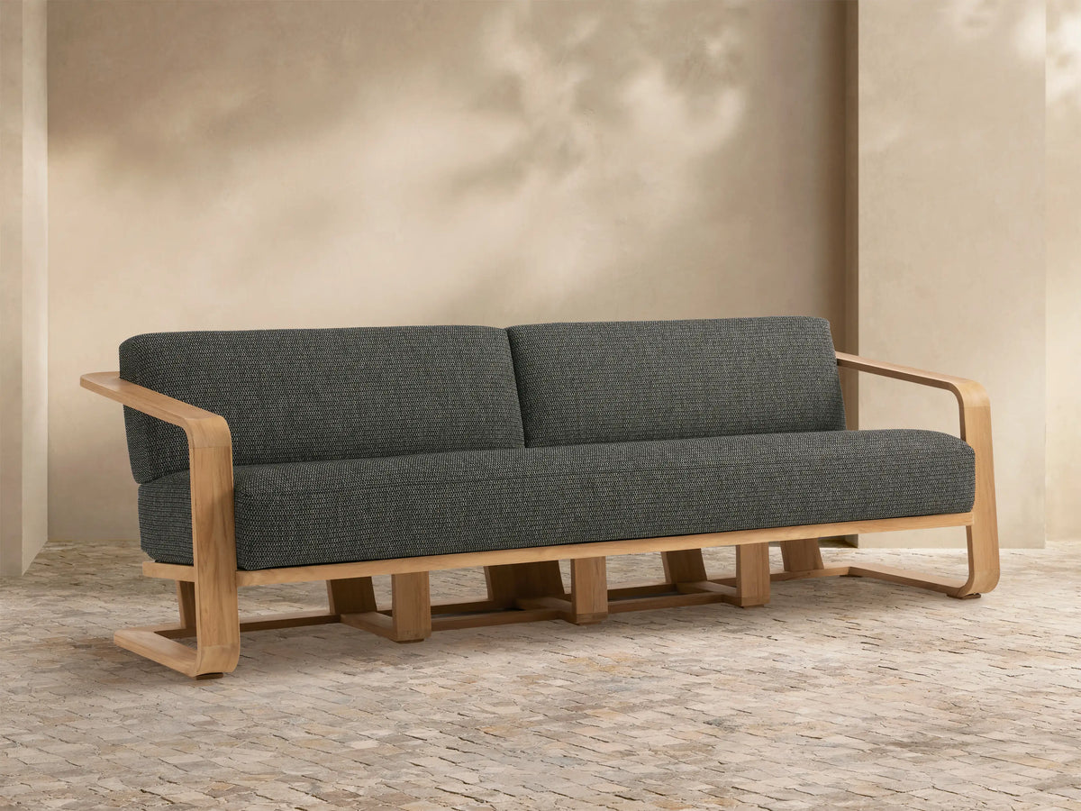 Sonoma Outdoor Teak Sofa: modern, light teak frame with dark gray cushions, two seats. Shown angled in an outdoor patio.