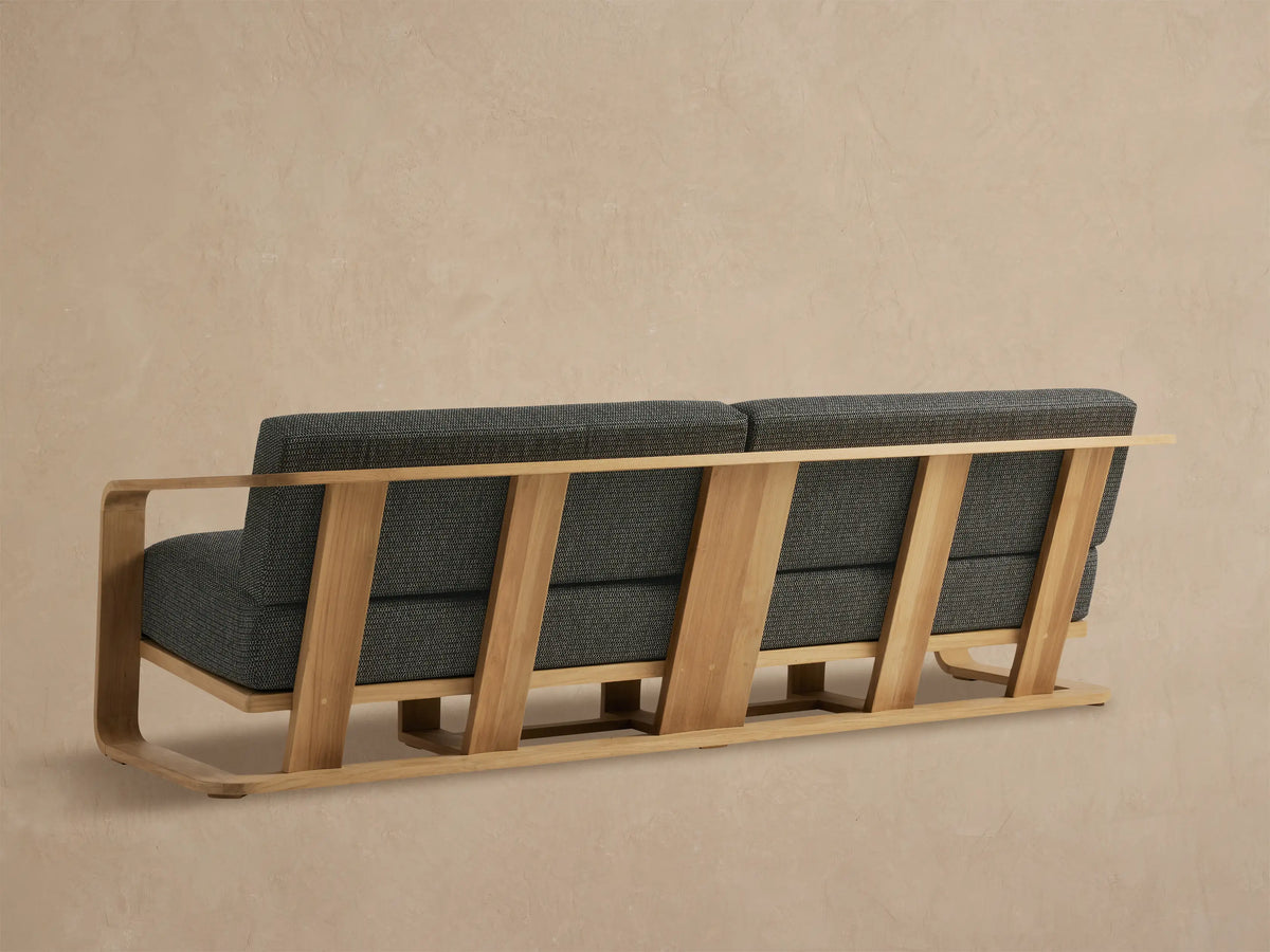 Sonoma Outdoor Teak Sofa close-up: smooth light teak armrest, dark grey marled fabric cushions, angled against beige.