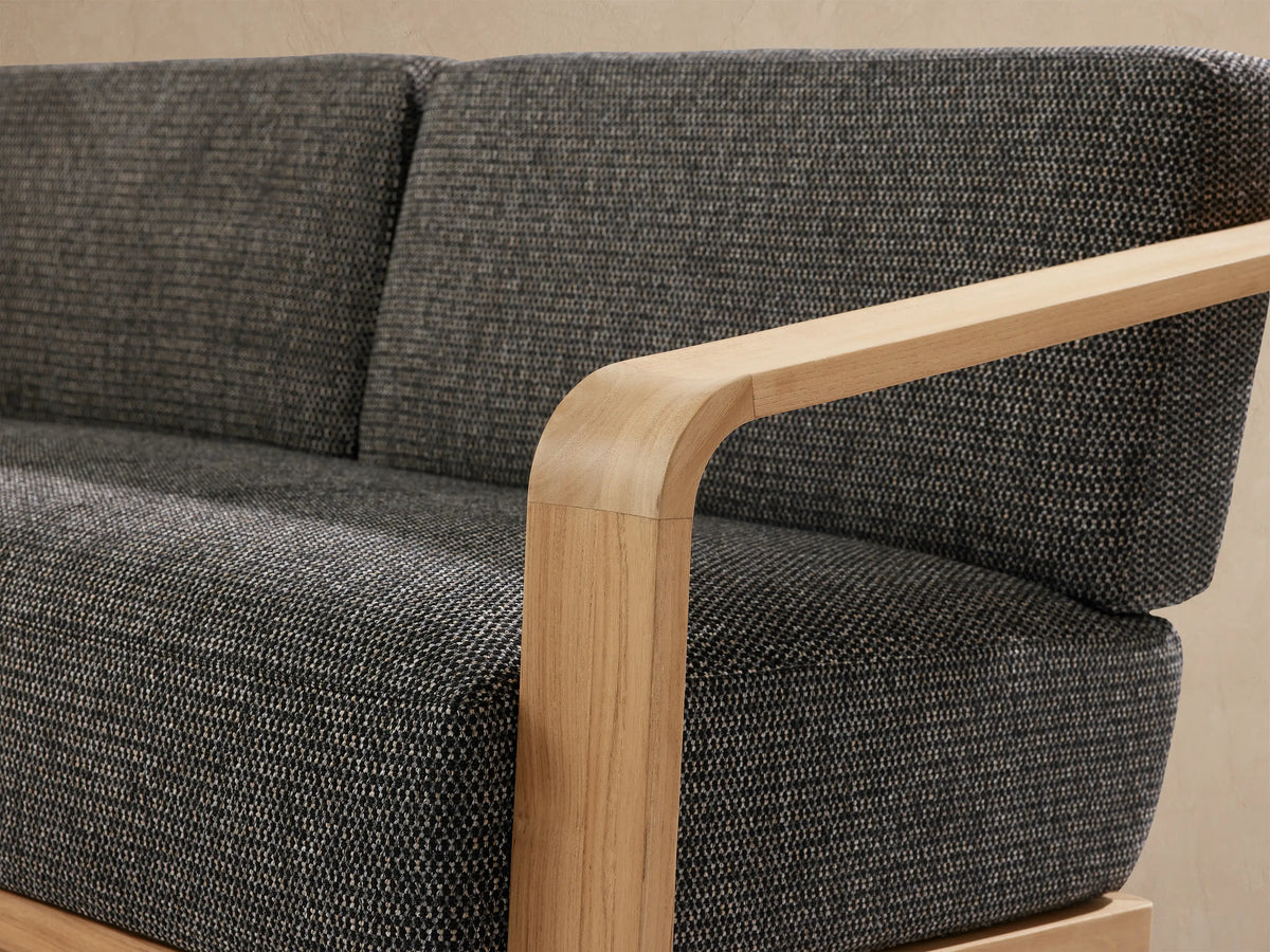 Sonoma Outdoor Teak Sofa, light natural teak frame with curved arms, dark gray textured cushions, presented slightly angled.