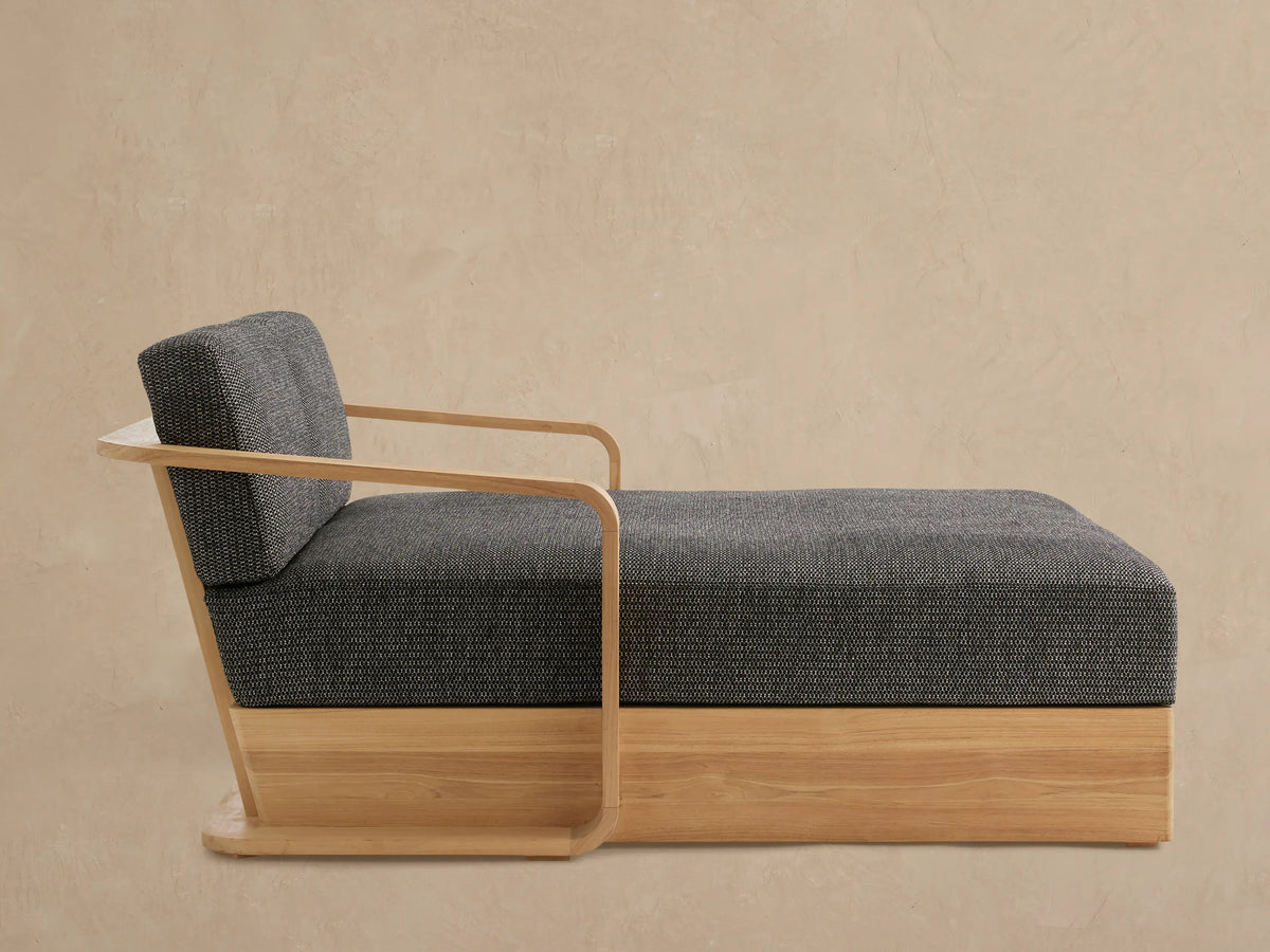 Sonoma Outdoor Teak Daybed with dark gray textured cushions, light natural teak wood frame, and curved armrests, angled indoors.