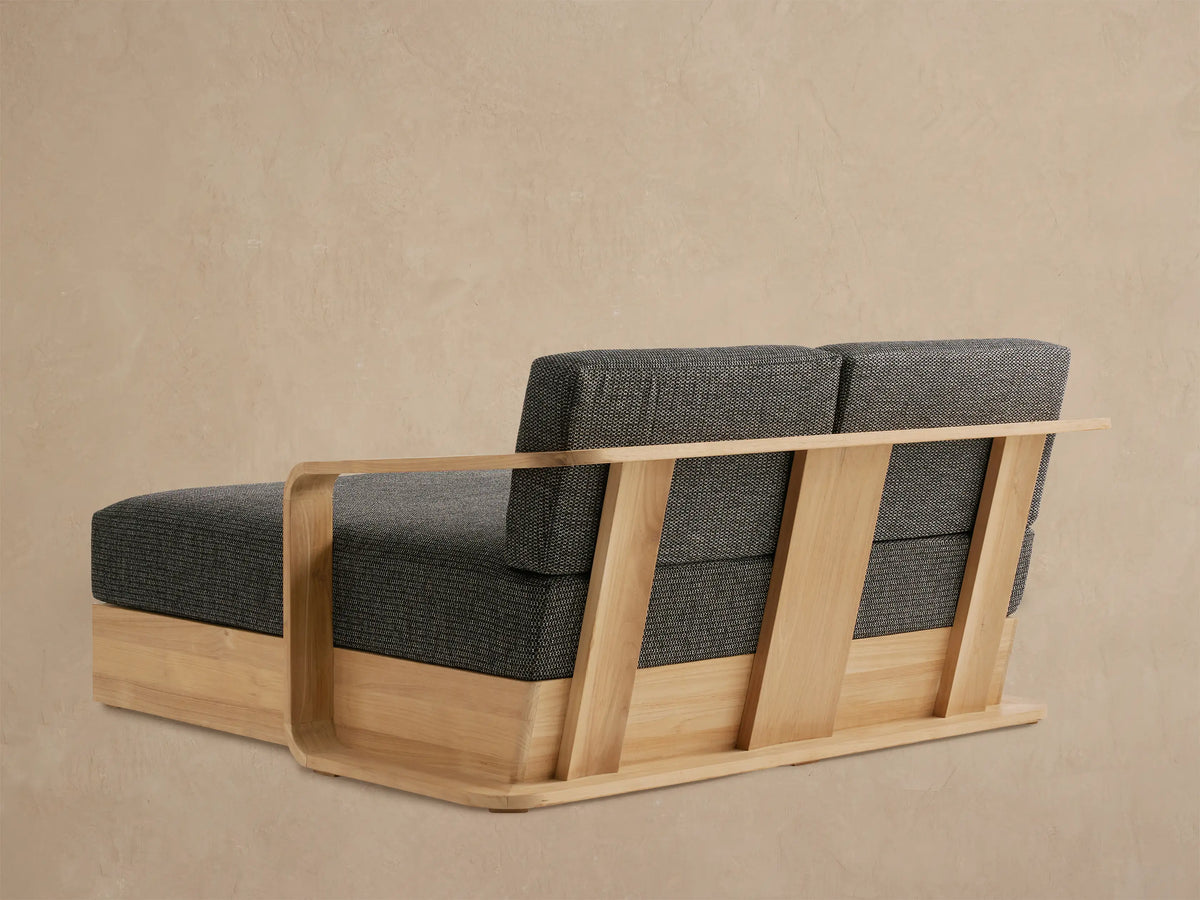 Angled close-up of Sonoma Teak Daybed armrest & cushions. Light wood frame with rounded edges, dark gray textured fabric.