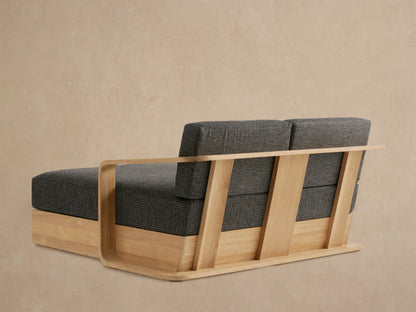 Angled close-up of Sonoma Teak Daybed armrest & cushions. Light wood frame with rounded edges, dark gray textured fabric.