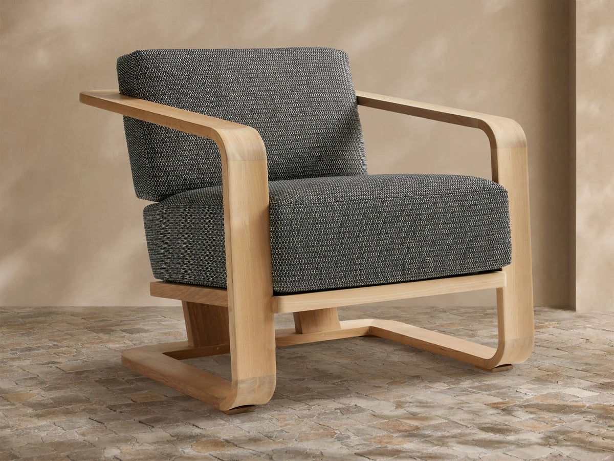 Sonoma Outdoor Teak Lounge Chair with light wood frame and dark gray, textured cushions, viewed from front-left on a tiled floor.