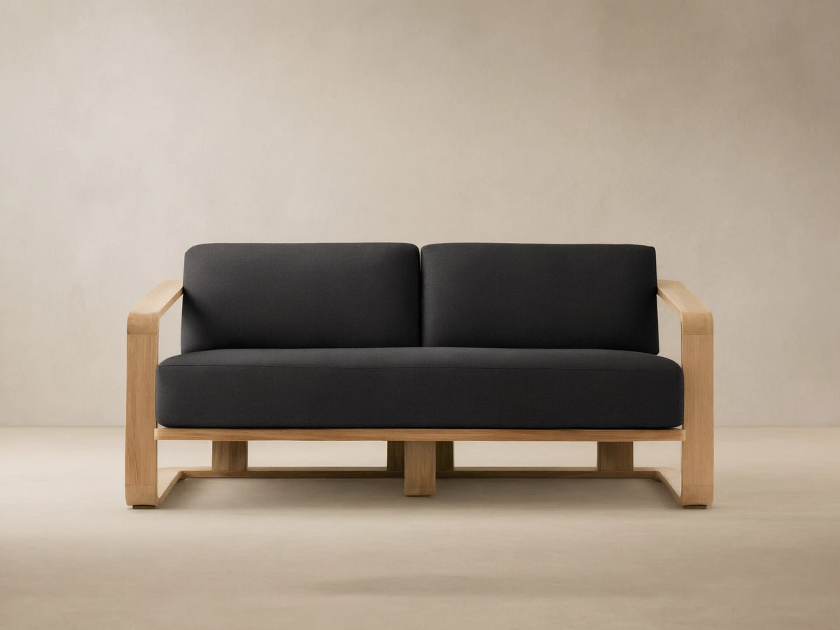 Front view of Sonoma Outdoor Teak Sofa with light teak frame & dark gray cushions, on stone floor before a beige wall.