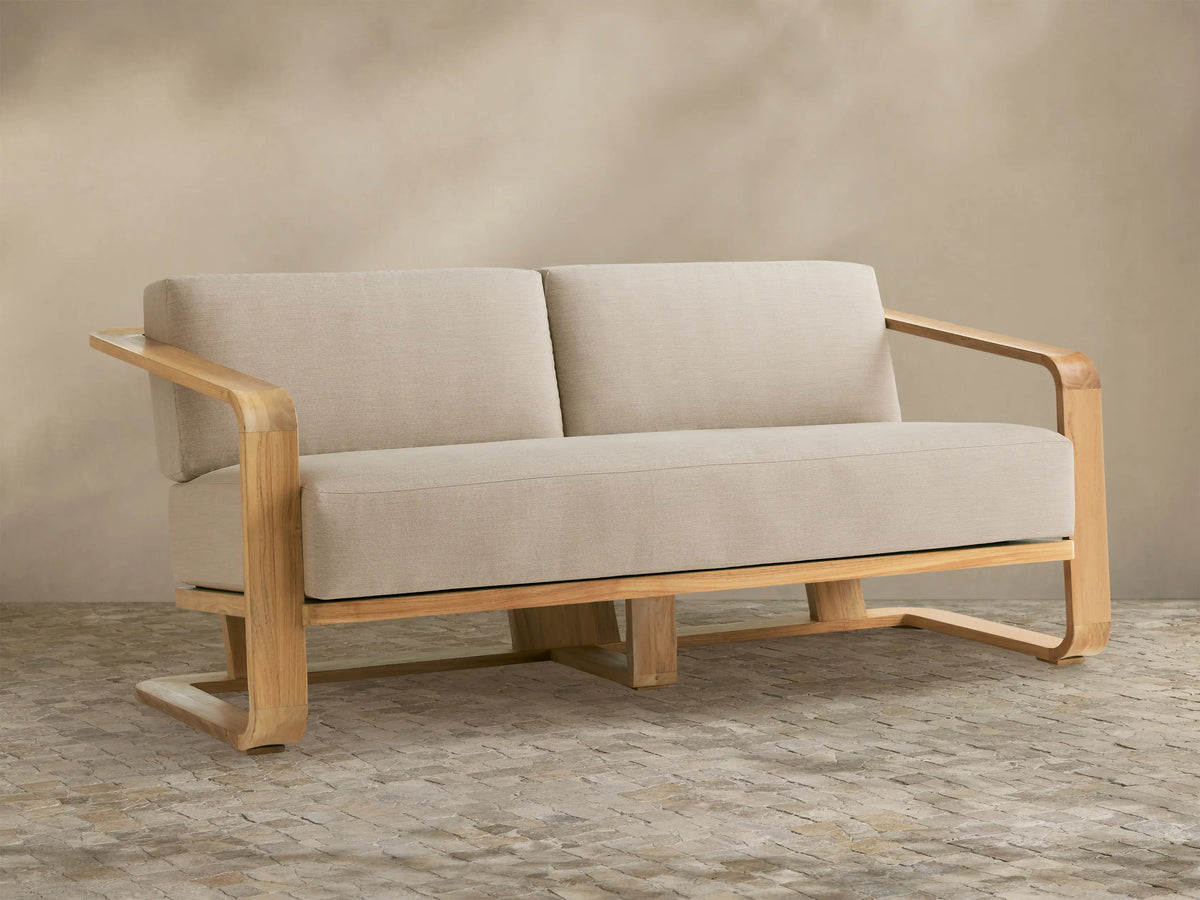 Sonoma Outdoor Teak Sofa, natural teak frame with wide arms, two light beige seat & back cushions, on a stone patio.