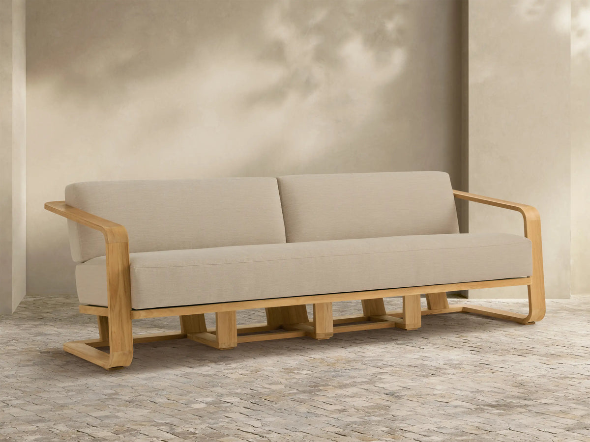 Sonoma Outdoor Teak Sofa with natural teak wood frame and light beige cushions, sitting on a textured patio against a shaded wall.