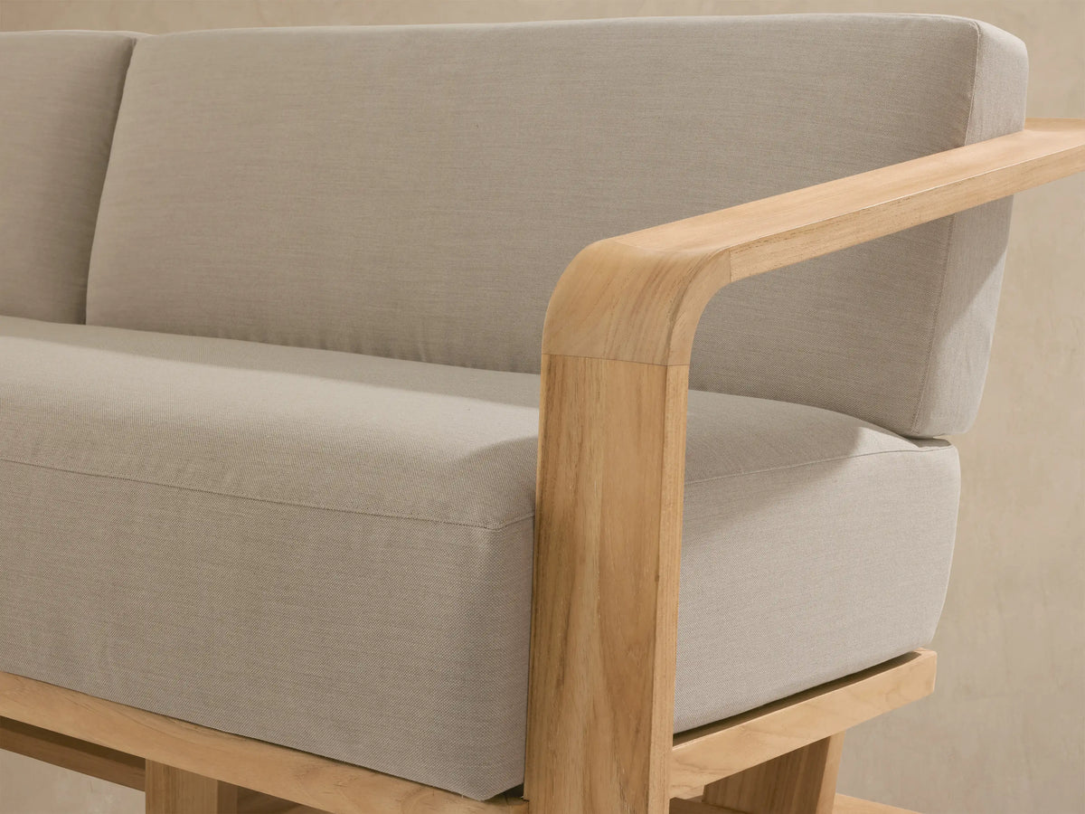 Sonoma Outdoor Teak Sofa with natural golden teak frame and light beige cushions, angled on an outdoor patio with dappled light.