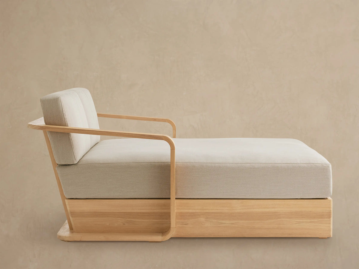Sonoma Outdoor Teak Daybed with natural teak wood base & curved armrests, light beige cushions, on a neutral paved patio.