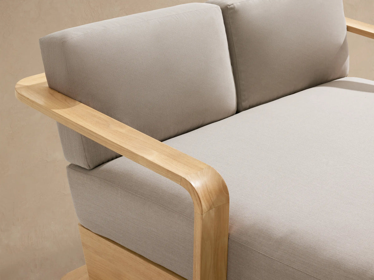 Sonoma Outdoor Teak Daybed, light natural teak frame with curved armrests, and light beige cushions, shown against a tan wall.