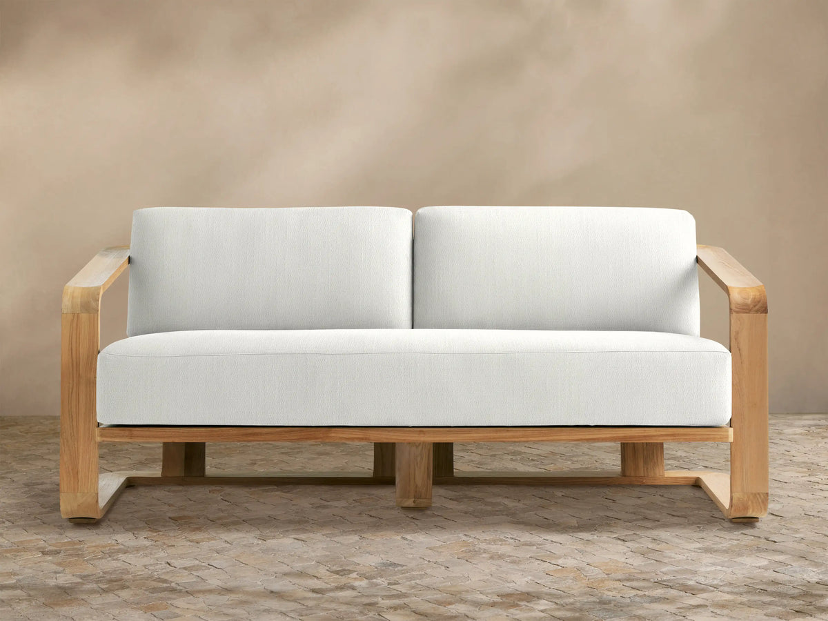 Sonoma Outdoor Teak Sofa. Light teak wood frame with two plump white cushions against a tan wall and stone-patterned floor.