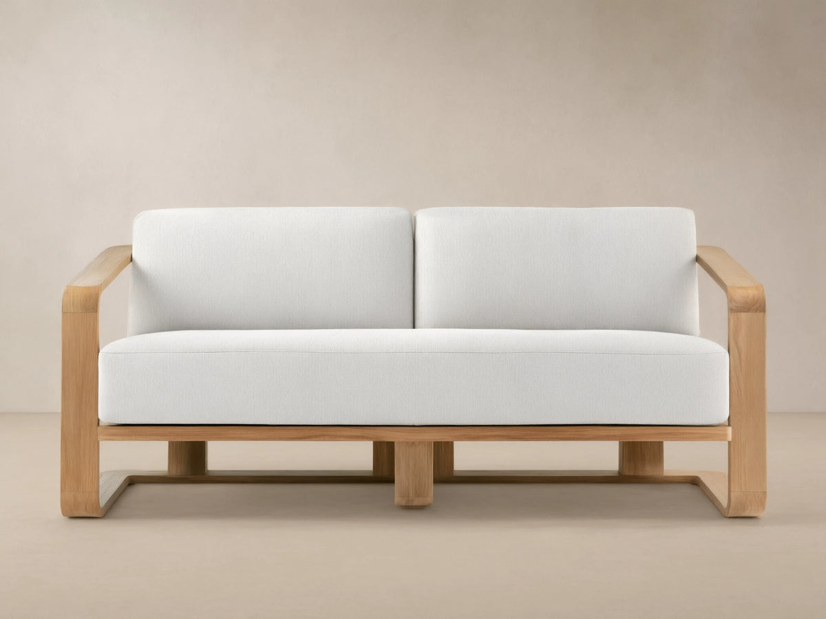 Sonoma Outdoor Teak Sofa. Light teak wood frame with two plump white cushions against a tan wall and stone-patterned floor.