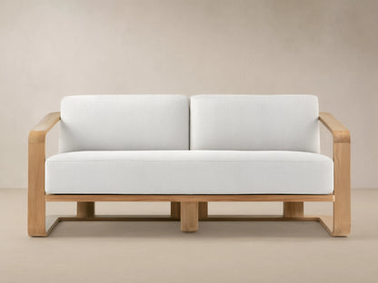 Sonoma Outdoor Teak Sofa. Light teak wood frame with two plump white cushions against a tan wall and stone-patterned floor.