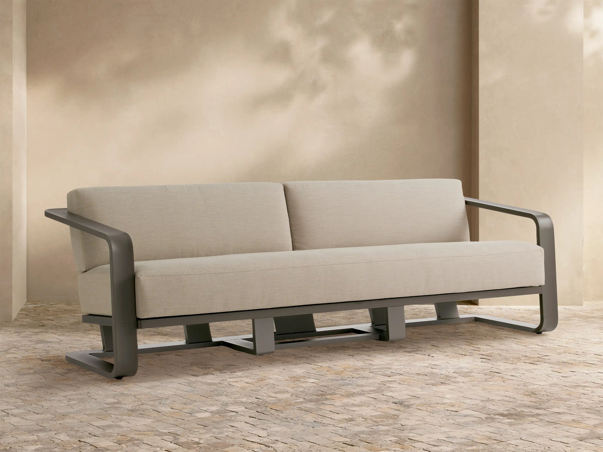 Sonoma Outdoor Aluminum Sofa with dark gray frame and light beige cushions, front view, on a light stone patio.