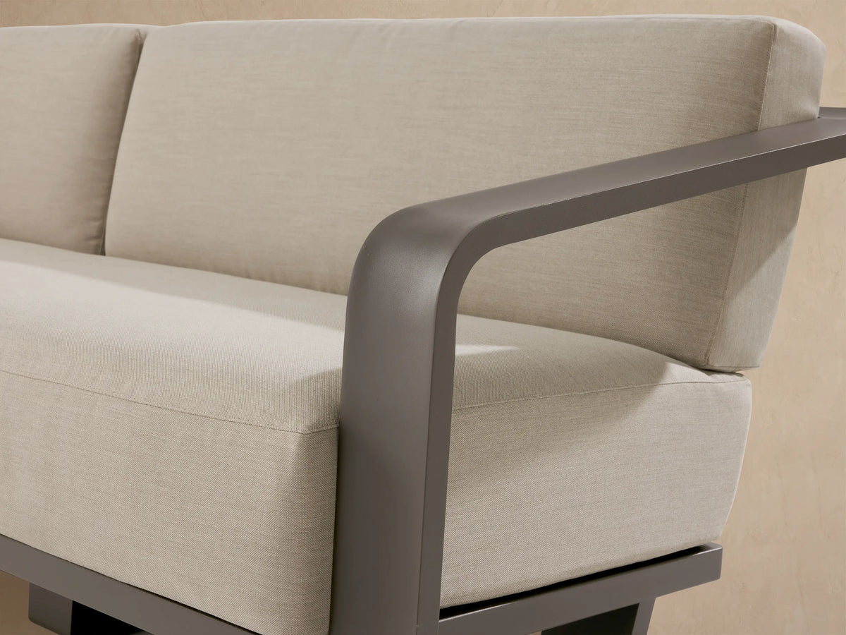 Sonoma Outdoor Aluminum Sofa: Dark gray frame, light beige cushions, on a light stone patio against a neutral stucco wall.