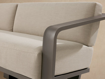 Sonoma Outdoor Aluminum Sofa: Dark gray frame, light beige cushions, on a light stone patio against a neutral stucco wall.