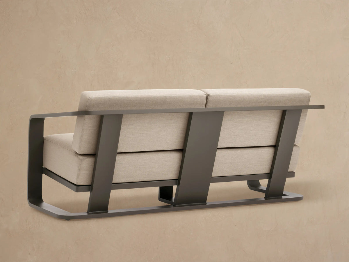 Close-up of Sonoma Outdoor Aluminum Sofa. Sleek dark gray aluminum armrest curves over light beige fabric seat and back cushions.