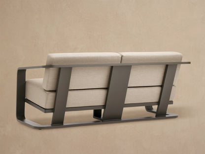 Close-up of Sonoma Outdoor Aluminum Sofa. Sleek dark gray aluminum armrest curves over light beige fabric seat and back cushions.
