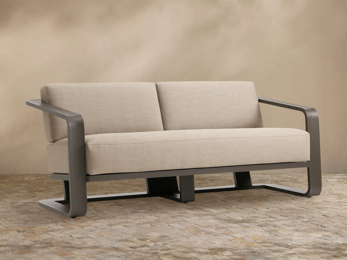 Sonoma Outdoor Aluminum Sofa, front. Dark gray frame, two light greige fabric cushions. Sits on stone patio against tan wall.