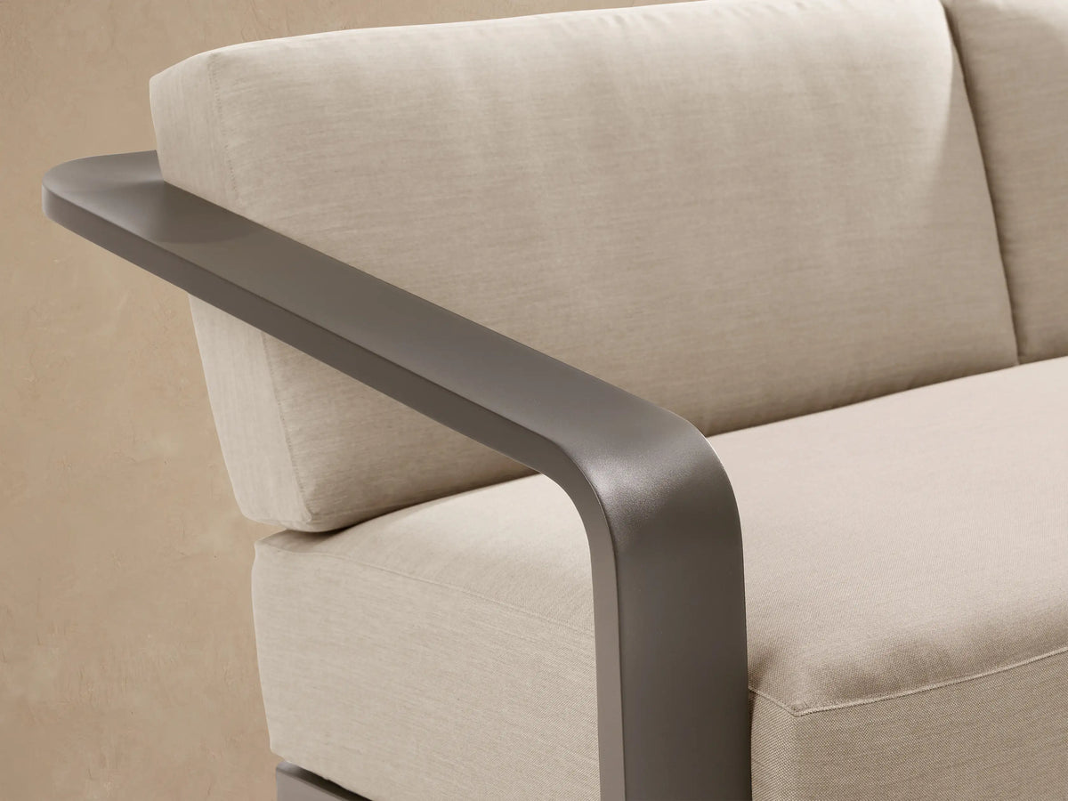 Sonoma Outdoor Aluminum Sofa, two-seater with dark gray frame, wide armrests, and light beige cushions on a stone patio.