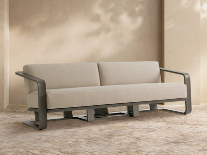 Sonoma Outdoor Aluminum Sofa with dark gray frame and light beige cushions, set on a stone patio against a light wall.