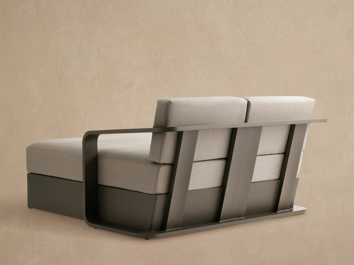 Angled close-up of Sonoma Outdoor Aluminum Daybed. Light gray cushions on a sleek, dark bronze aluminum frame with curved armrest.