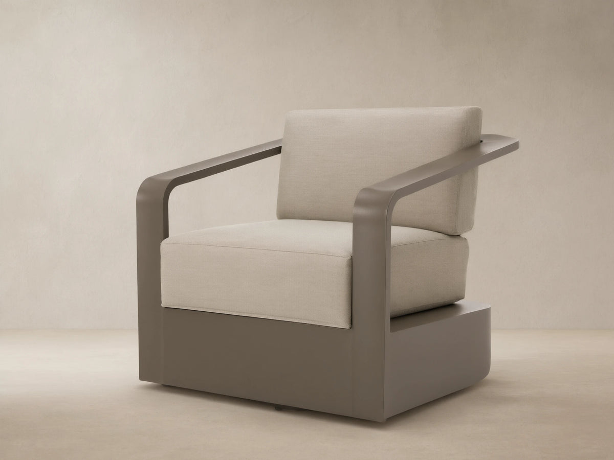 Sonoma Outdoor Aluminum Swivel Chair with sleek brown frame and thick, light beige cushions on a stone patio.