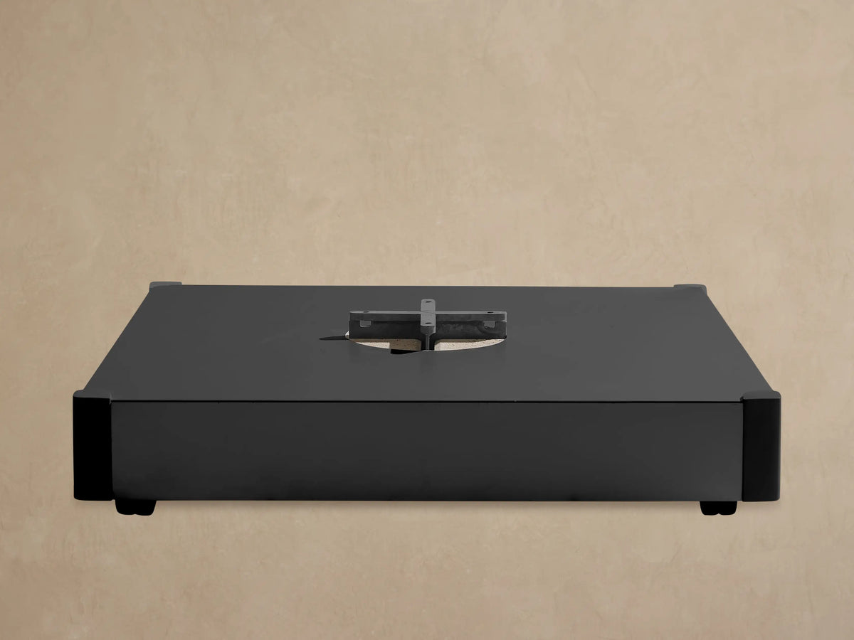 Black rectangular Spectra Outdoor Umbrella Base with a central silver cross-shaped mounting plate against a light wall.