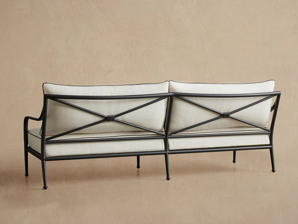 Surrey Outdoor Sofa, angled close-up view. Black aluminum frame with X-shaped back, light beige cushions with black piping.