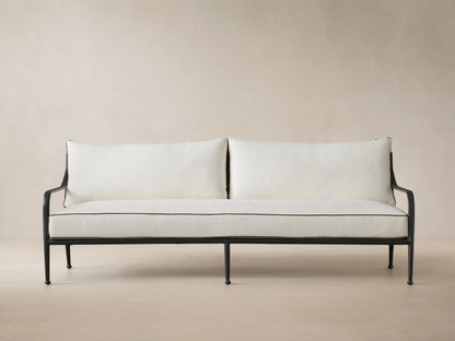 Surrey Outdoor Sofa: black metal frame, two white cushions with black piping, front view, against a beige wall.
