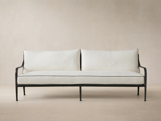 Surrey Outdoor Sofa: black metal frame, two white cushions with black piping, front view, against a beige wall.
