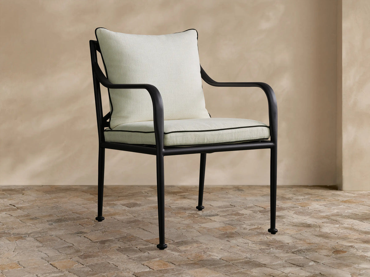 Surrey Outdoor Dining Arm Chair. Black powder-coated frame, light cream cushions with black piping, on a stone patio.