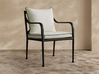 Surrey Outdoor Dining Arm Chair. Black powder-coated frame, light cream cushions with black piping, on a stone patio.