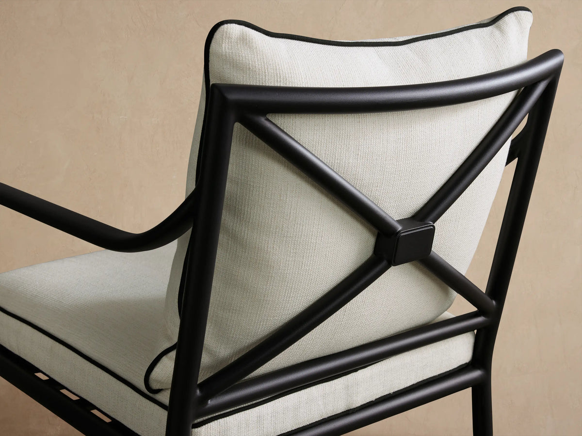Surrey Outdoor Dining Arm Chair, front, black aluminum frame, light cream cushions with black piping, on a textured stone floor.