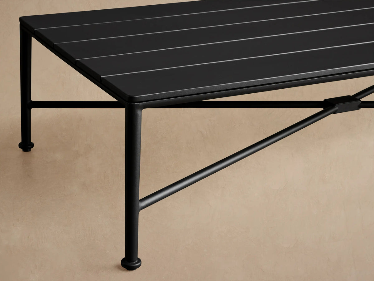 Black rectangular Surrey Outdoor Coffee Table with slatted top, sturdy aluminum frame, on a stone patio against a tan wall.