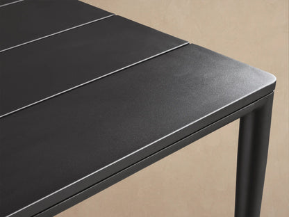 Rectangular black Surrey Outdoor Dining Table with umbrella hole on a light brown paved patio, against a textured beige wall.