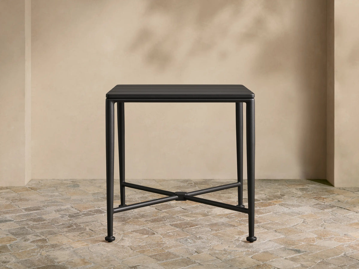 Matte black Surrey Outdoor End Table with square slatted top, sturdy legs, and X-cross support on a stone patio.