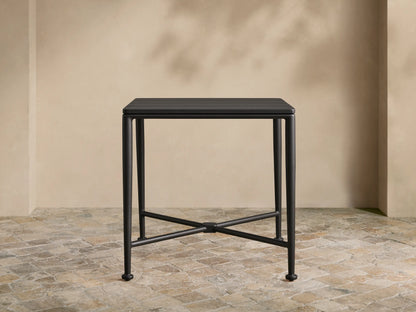 Matte black Surrey Outdoor End Table with square slatted top, sturdy legs, and X-cross support on a stone patio.