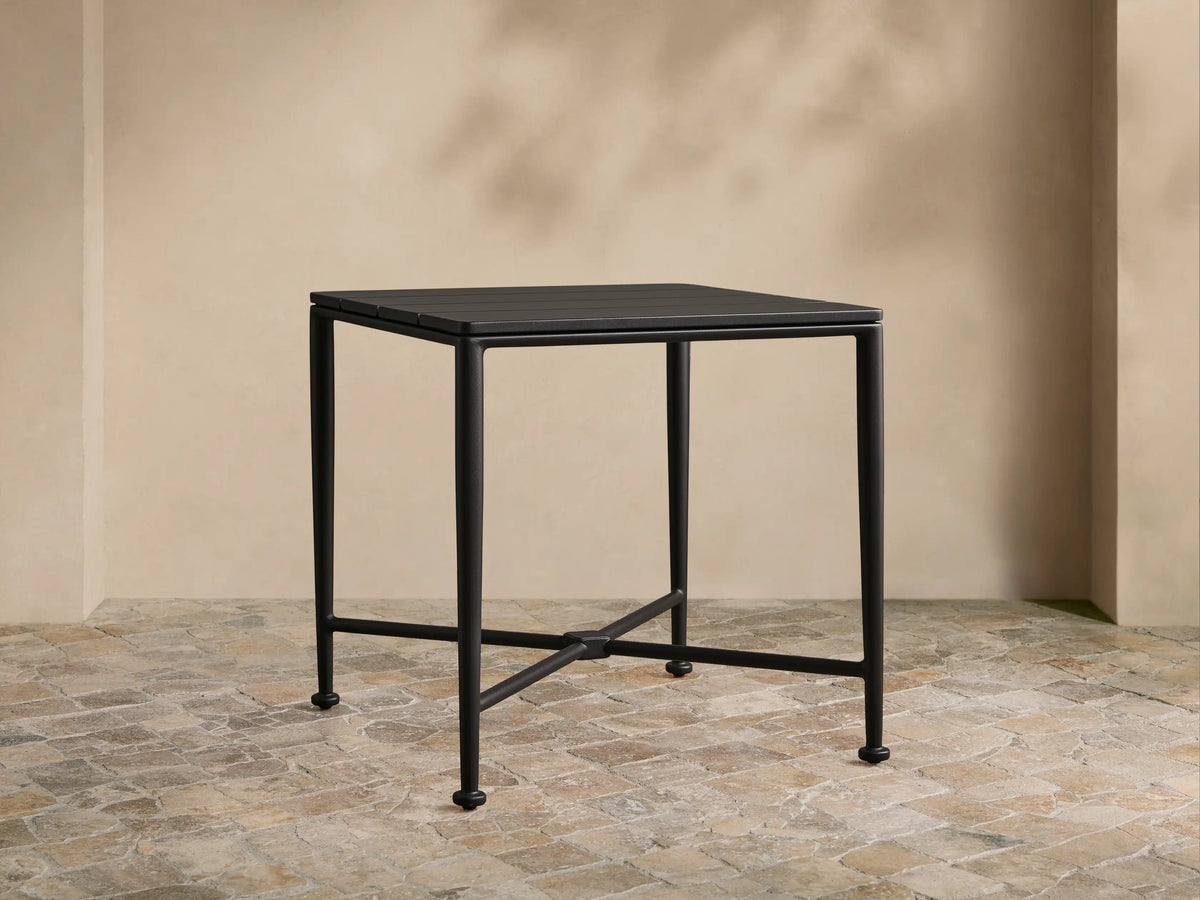 Matte black Surrey Outdoor End Table with square slatted top, sturdy legs, and X-cross support on a stone patio.