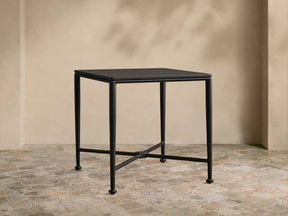 Matte black Surrey Outdoor End Table with square slatted top, sturdy legs, and X-cross support on a stone patio.