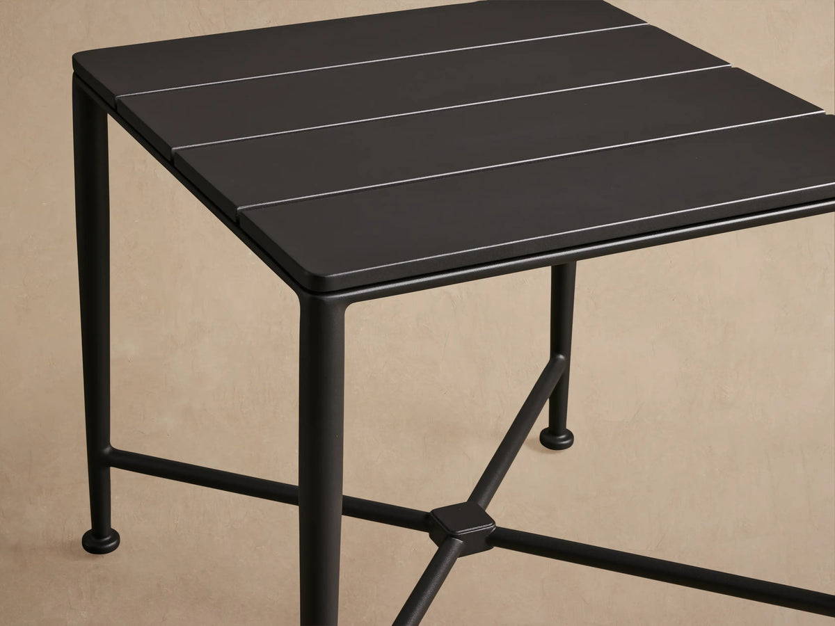 Surrey Outdoor End Table. Square black aluminum table with slatted top, four straight legs, on a stone patio.