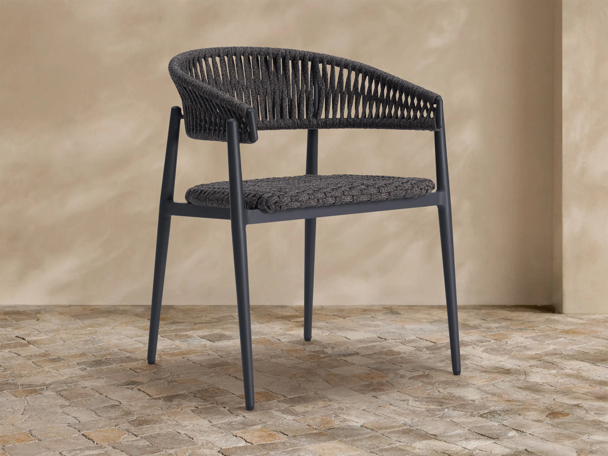 Talia Outdoor Dining Chair. Dark gray woven rope back and seat, sleek dark frame, on a textured stone floor.
