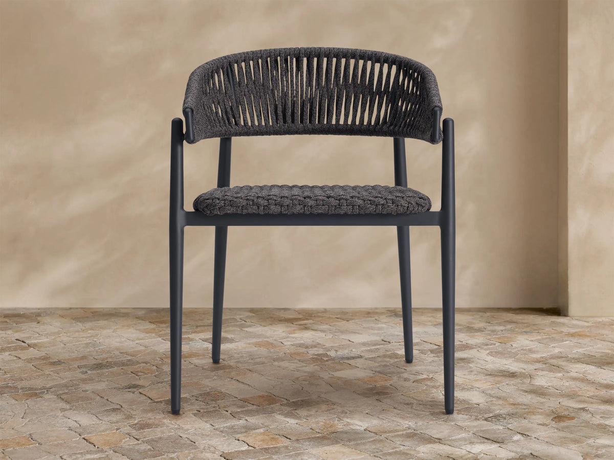 Talia Outdoor Dining Chair. Dark gray woven rope back and seat, sleek dark frame, on a textured stone floor.